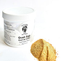 A white container of Grand Valley Quail Pet Treats quail egg food topper for dogs, placed beside a small pile of fine golden powder. The label highlights its nutritional benefits and use as a meal enhancer.